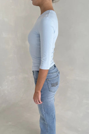 Model in profile wearing a light blue Esme High V 3/4 Sleeve Top paired with high-rise jeans and white sandals in a neutral studio.