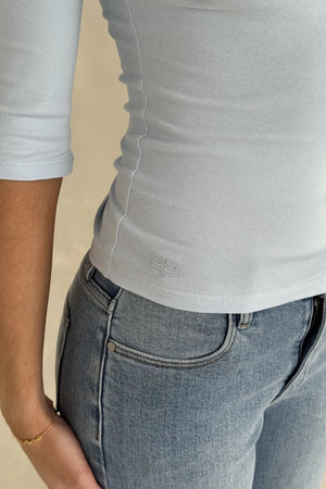 Model wears a light blue ribbed Esme High V 3/4 Sleeve Top with subtle 'SS' logo, paired with classic blue denim jeans.