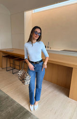 A woman in a light blue Esme High V 3/4 Sleeve Top pairs it with blue jeans, white heels, and a leopard print handbag in a modern kitchen.