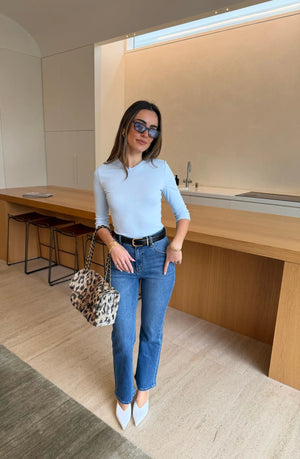 A woman in a light blue Esme High V 3/4 Sleeve Top pairs it with blue jeans, white heels, and a leopard print handbag in a modern kitchen.
