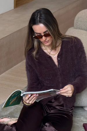 A woman wearing a dark purple Kai Cardigan Chocolate and chocolate satin trousers sits on a rug reading a magazine in a modern room.