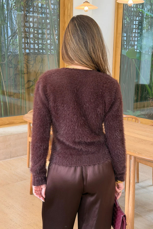 A woman wears the Kai Cardigan Chocolate with satin trousers and holds a woven plum handbag in a bright, modern room.