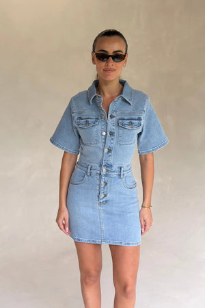 Light blue denim mini dress with short sleeves and a fitted silhouette, styled with front buttons and a casual look.