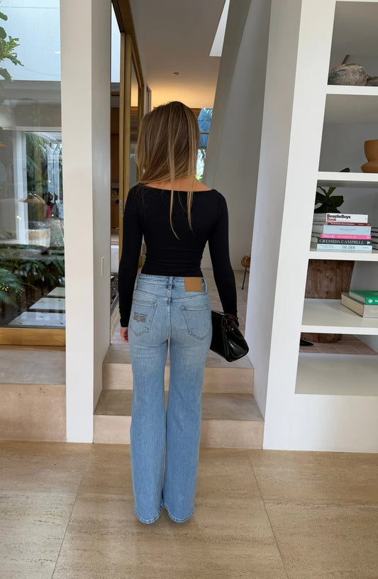 A woman in a black top wears Kendall Mid Rise Flare Jeans in mid wash, standing in a bright, minimalist room with white walls and beige floors.