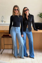 Two women wear Kendall Mid Rise Flare Jeans in mid wash with black tops, sunglasses, and heels in a modern, sunlit kitchen setting.