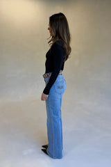 A woman in a black long-sleeve top and Kendall Mid Rise Flare Jeans with a snakeskin belt and black heels stands in profile.