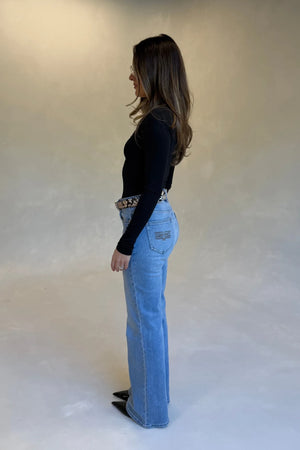 A woman in a black long-sleeve top and Kendall Mid Rise Flare Jeans with a snakeskin belt and black heels stands in profile.