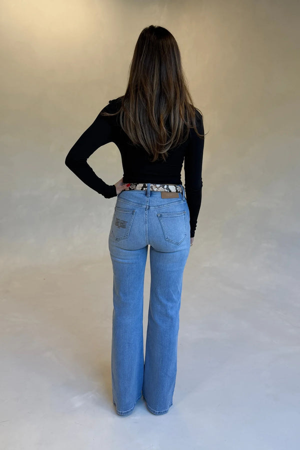 A woman wears Kendall Mid Rise Flare Jeans in mid wash with a snakeskin belt and black fitted top, shown from behind.