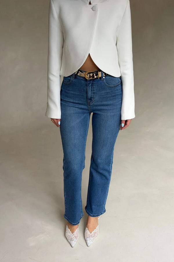 A person wears Kennedy Crop Jean Indigo with a white blazer, snakeskin belt, and white lace pointed-toe heels against a grey background.