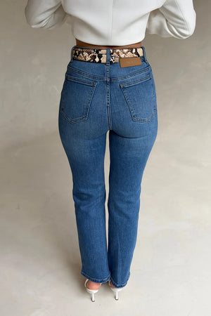 Woman wearing high-waisted Kennedy Crop Jean Indigo with a snakeskin belt, white long-sleeve top, and white stiletto heels.
