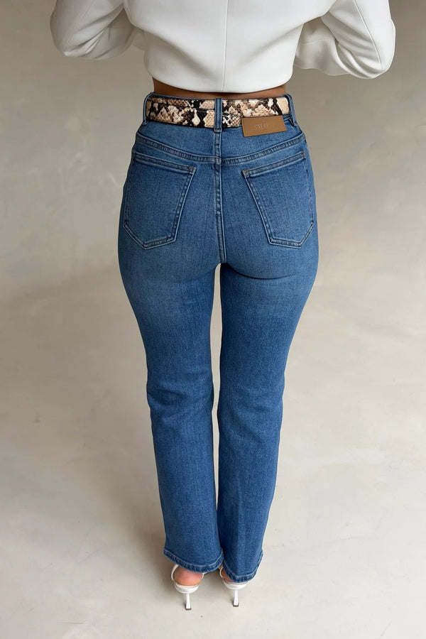 Woman wearing high-waisted Kennedy Crop Jean Indigo with a snakeskin belt, white long-sleeve top, and white stiletto heels.