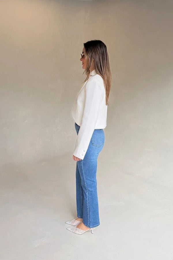 A woman in profile wears Kennedy Crop Jean Indigo with a white blazer and lace kitten heels against a minimalist beige and grey studio backdrop.