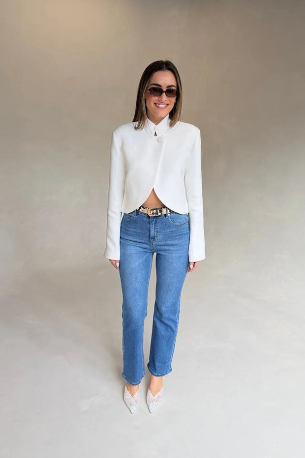 A woman wears Kennedy Crop Jean Indigo with a white cropped jacket, leopard-print belt, white heels, and brown sunglasses against a grey backdrop.