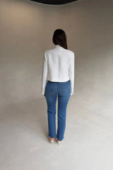 A woman wears a white cropped blazer and high-waisted Kennedy Crop Jean Indigo with light heeled sandals against a gray backdrop.