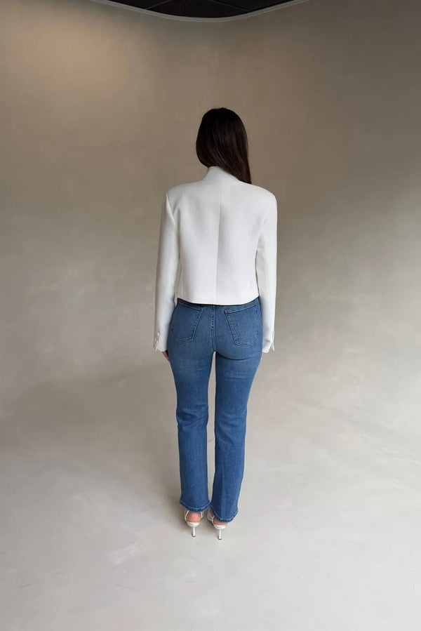 A woman wears a white cropped blazer and high-waisted Kennedy Crop Jean Indigo with light heeled sandals against a gray backdrop.