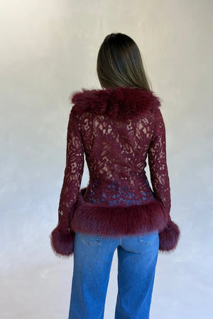 A woman wears a burgundy lace long-sleeve top with faux fur trim on collar, cuffs, and hem, paired with blue jeans.