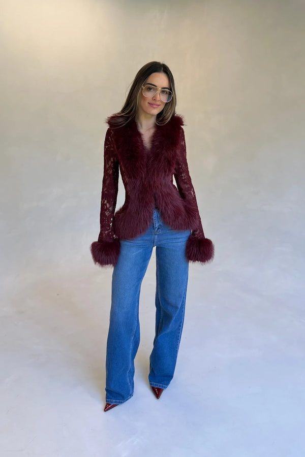 A woman wears a burgundy lace fur trim top with blue jeans, burgundy heels, and clear-framed glasses against a grey background.