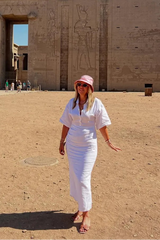 Woman in a white outfit and pink hat standing in front of an ancient temple with hieroglyphics.