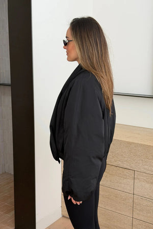 A young woman in a Maddison Shawl Collar Oversized Jacket Black and designer sunglasses poses against tan tiled walls in a minimalist setting.