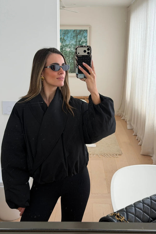 A woman wears the Maddison Shawl Collar Oversized Jacket Black with black leggings, gold hoop earrings, and dark sunglasses in a minimalist room.
