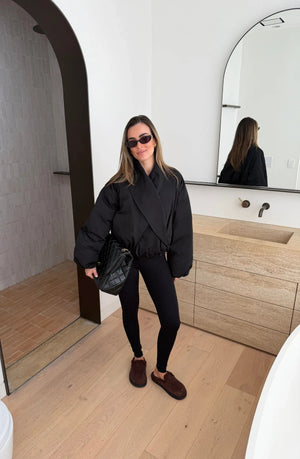 A woman wearing a black Maddison Shawl Collar Oversized Jacket poses in a modern bathroom with arched doorways and light hardwood floors.