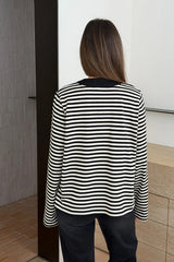 Person wearing a black and white striped Millie Relaxed Long Sleeve Tee with dark trousers, standing in a minimalist interior.