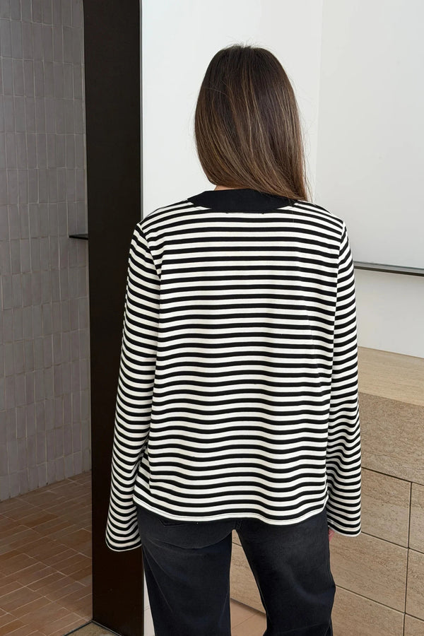Person wearing a black and white striped Millie Relaxed Long Sleeve Tee with dark trousers, standing in a minimalist interior.