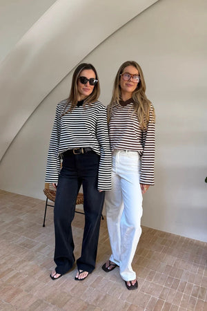 Two women pose in a bright, minimalist room wearing Millie Relaxed Long Sleeve Tees in black/white and brown/white stripes.