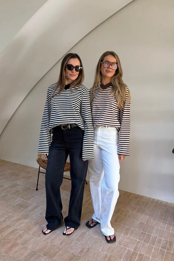 Two women pose in a bright, minimalist room wearing Millie Relaxed Long Sleeve Tees in black/white and brown/white stripes.