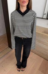 A woman wears the Millie Relaxed Long Sleeve Tee in black and white stripes with black jeans and gold accessories in a minimalist room.