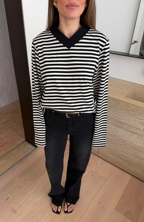 A woman wears the Millie Relaxed Long Sleeve Tee in black and white stripes with black jeans and gold accessories in a minimalist room.