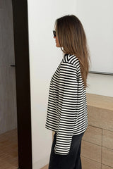 A woman in a Millie Relaxed Long Sleeve Tee with black and white stripes stands sideways in a minimalist room wearing dark pants and Gucci sunglasses.