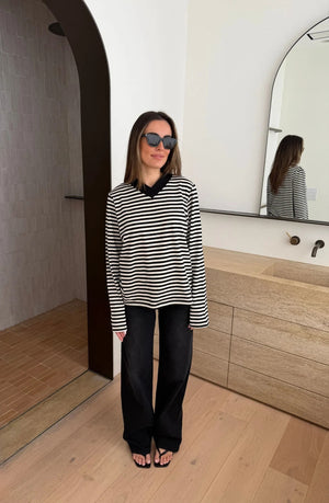 A woman wears the Millie Relaxed Long Sleeve Tee in black and white stripes with black trousers and sunglasses in a minimalist bathroom.