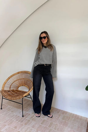 A woman wears the Millie Relaxed Long Sleeve Tee Chocolate Stripe with wide-leg black jeans and sunglasses in a minimalist room.