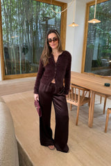 A woman wears chocolate satin wide-leg pants with a fuzzy cardigan and sunglasses in a bright, modern room overlooking a bamboo garden.