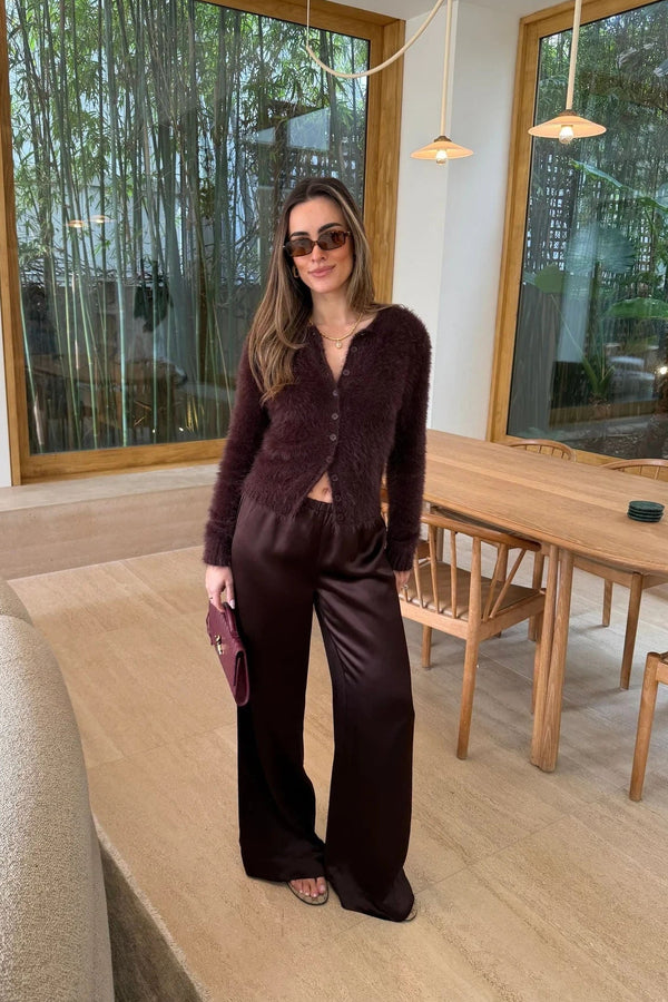 A woman wears chocolate satin wide-leg pants with a fuzzy cardigan and sunglasses in a bright, modern room overlooking a bamboo garden.