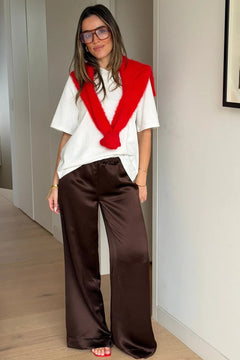 A woman wears Naomi Relaxed Satin Pant Chocolate with a white tee, red sweater draped over shoulders, tortoiseshell glasses, and red sandals.