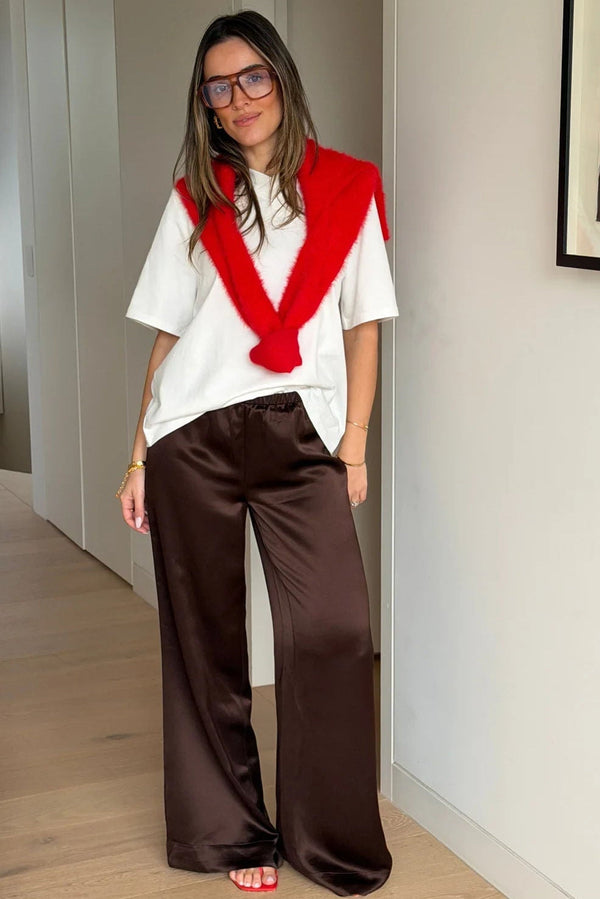 A woman wears Naomi Relaxed Satin Pant Chocolate with a white tee, red sweater draped over shoulders, tortoiseshell glasses, and red sandals.