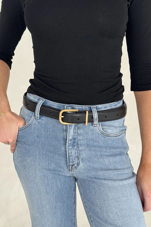 A slim Renee Leather Belt Black with a gold buckle cinches light-wash jeans paired with a fitted black top and gold bracelet.