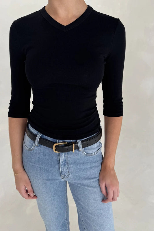 A woman wears a Renee Leather Belt Black with a fitted black ribbed top and light-wash blue jeans against a light-grey background.