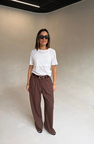 A woman wears a Renee Leather Belt Chocolate with dark brown pinstriped trousers, a white layered tee, gold bracelets, and Gucci sunglasses.