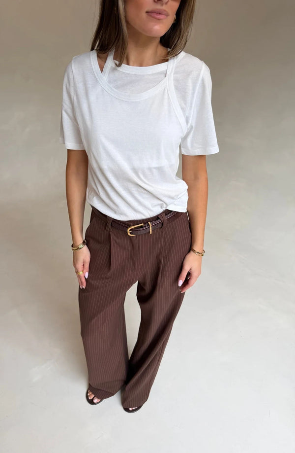 A woman wears the Renee Leather Belt Chocolate with a layered white top and dark brown pinstriped trousers, accessorized with gold jewelry.