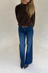 A woman wears a Renee Leather Belt Chocolate with a dark brown sweater, bootcut blue jeans, and brown suede heels against a grey backdrop.