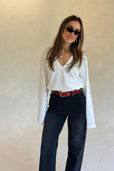 A woman wears a Renee Leather Belt Red Croc with a white v-neck top, black jeans, gold hoop earrings, pearl necklace, and dark sunglasses.