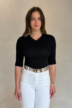 A woman wears the Renee Leather Belt Snake with a gold buckle, styled over white jeans and a fitted black top in a minimalist setting.