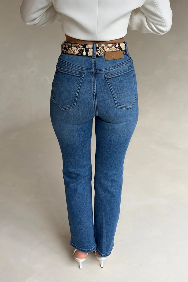 Woman wearing high-waisted blue jeans with the Renee Leather Belt Snake, a snakeskin-patterned belt, paired with a white top and heels.