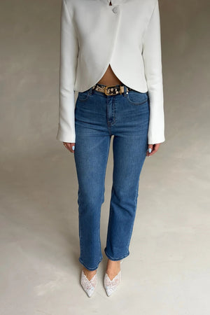 A person wears a Renee Leather Belt Snake with a white blazer, blue jeans, and white lace pointed-toe heels against a grey background.