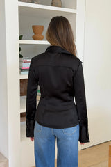 A woman stands facing away wearing a black satin Tara long sleeve shirt paired with classic blue denim jeans in a bright, minimalist room.