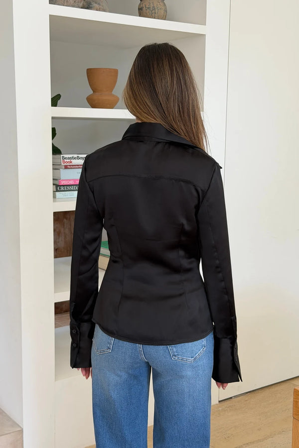 A woman stands facing away wearing a black satin Tara long sleeve shirt paired with classic blue denim jeans in a bright, minimalist room.