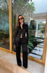 A woman wears the Tara Satin Long Sleeve Shirt Black paired with wide-leg trousers, a snakeskin belt, gold earrings, and a black quilted Chanel bag.